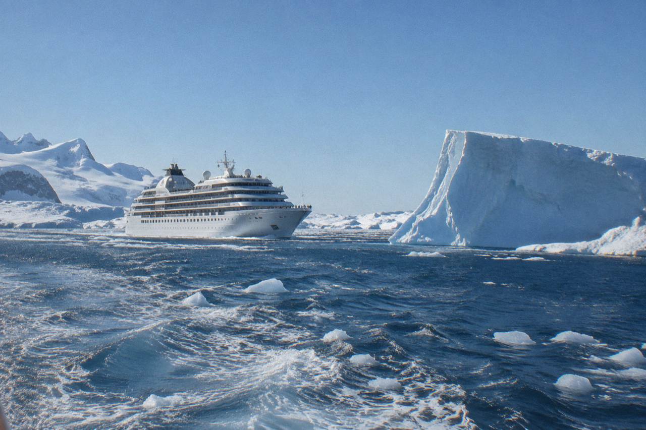 A large white cruise ship cruising past icebergs and snowy Antarctic coastline