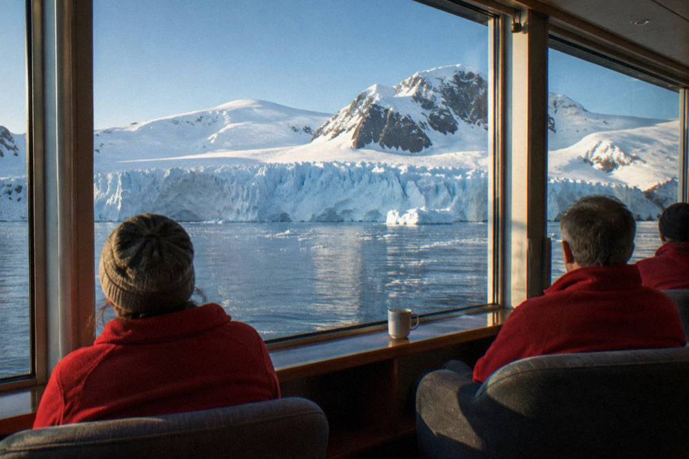 Ship lounge with panoramic windows and guests seated looking out at icebergs