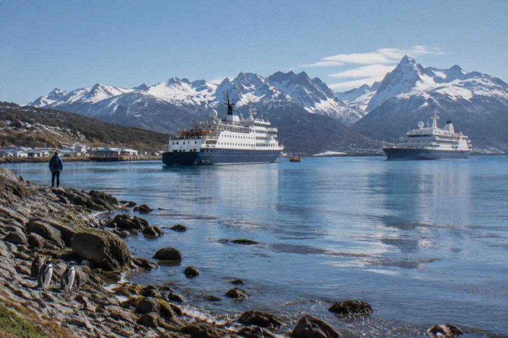 Two expedition ships at anchor in a calm Antarctic bay with mountains behind
