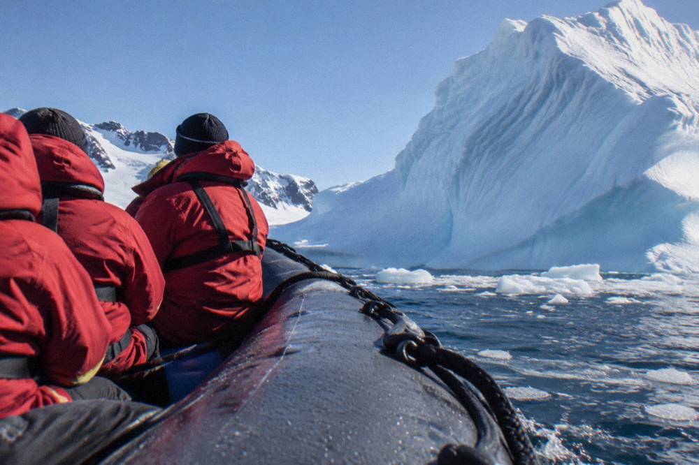 Zodiac cruising past a tall iceberg near the Antarctic Peninsula shoreline