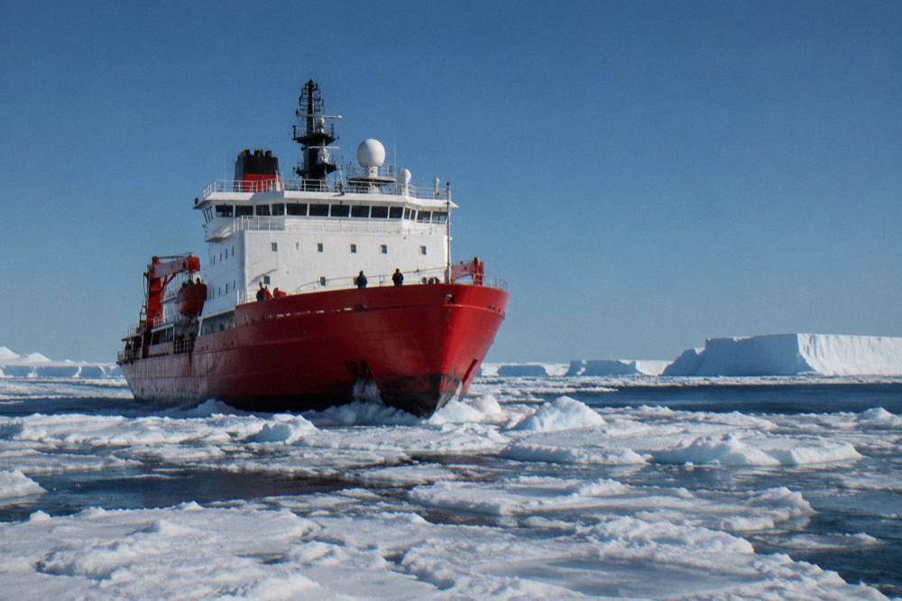 A red and white expedition ship moving through loose brash ice under a grey polar sky