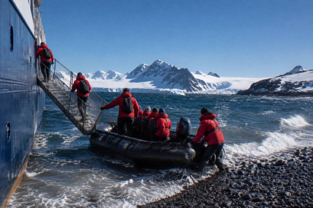 Passengers in red parkas boarding a Zodiac from a gangway beside an ice-strengthened ship hull