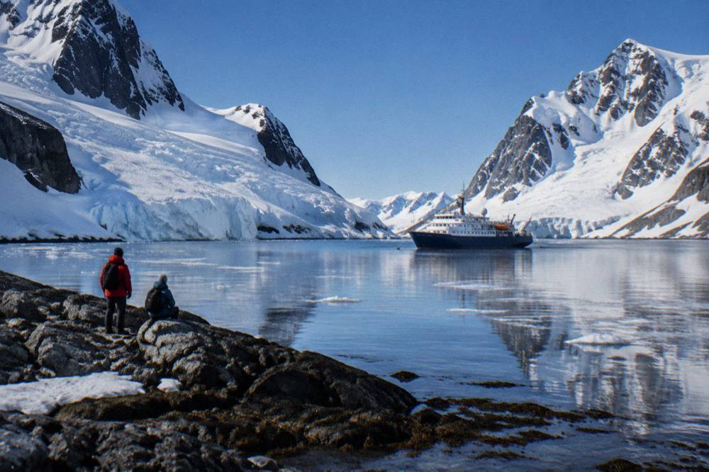 An expedition ship anchored in a calm bay with steep glaciers running down to black rock shoreline