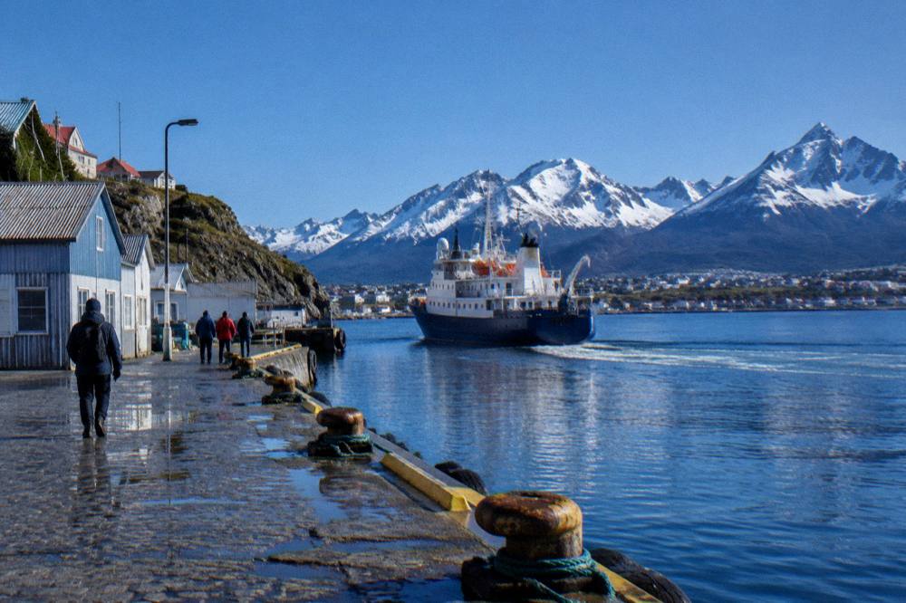 A small expedition ship leaving a grey Ushuaia harbour with snow-capped mountains behind the waterfront buildings