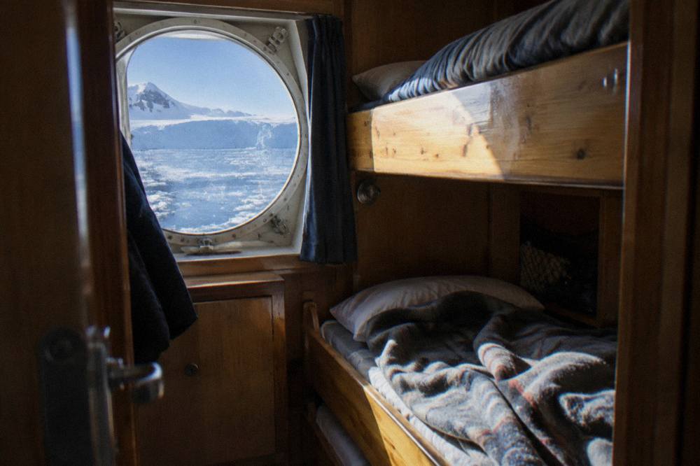 A small ship cabin with twin bunks beside a round porthole showing icy water