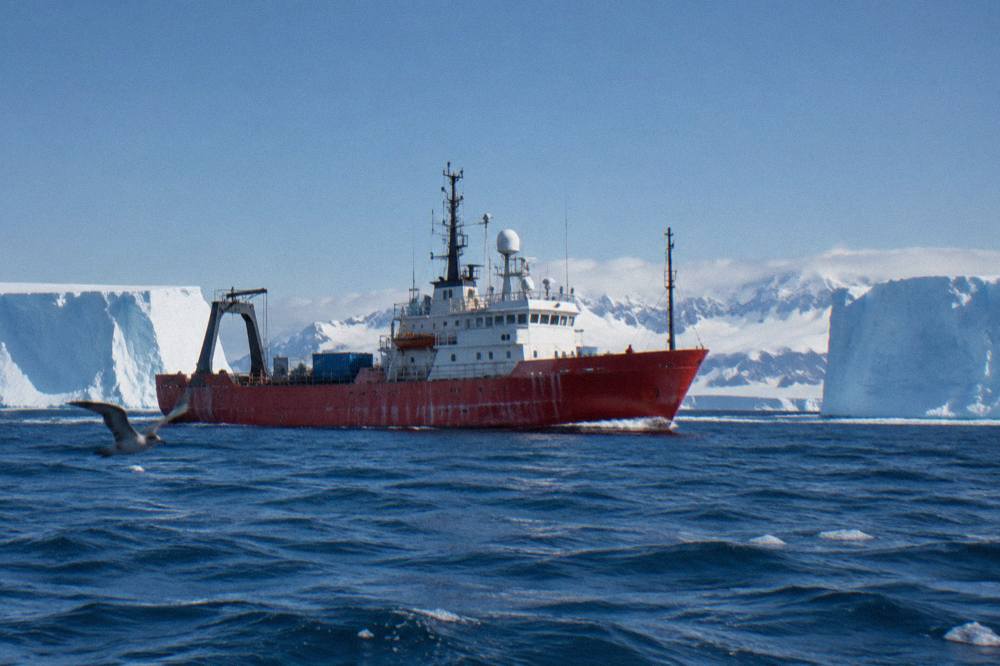 Small red-hulled expedition ship moving through calm polar water with icebergs