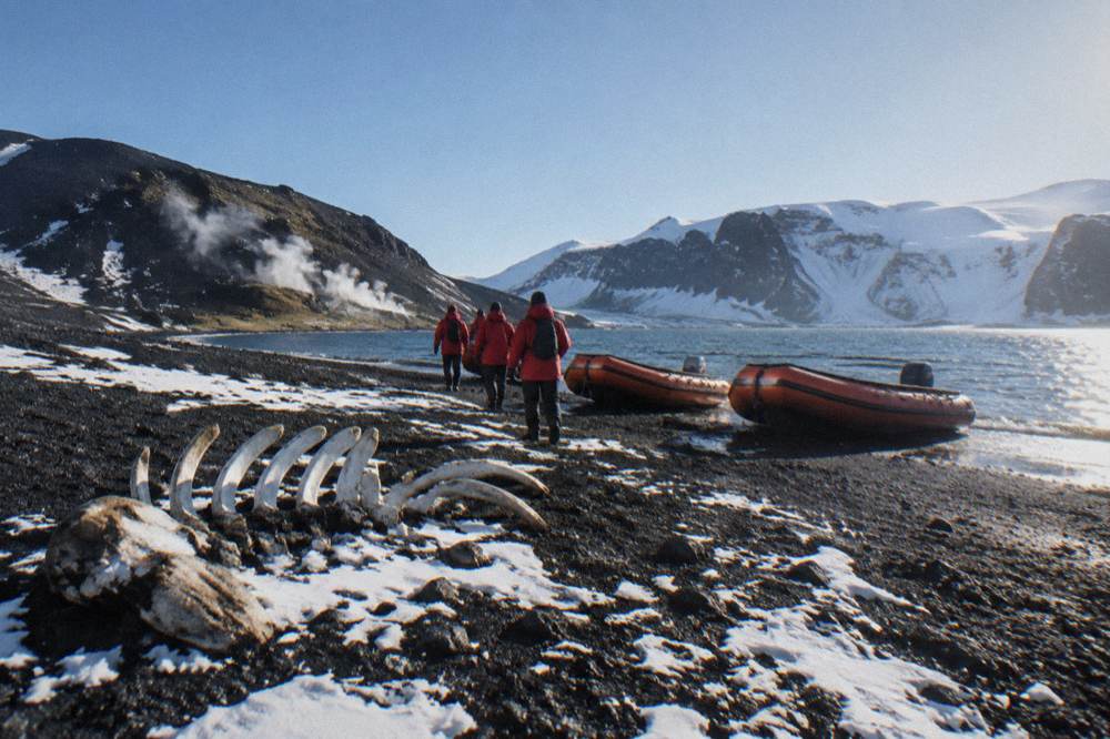 Travellers in red parkas standing on a snowy Antarctic beach beside orange Zodiac boats