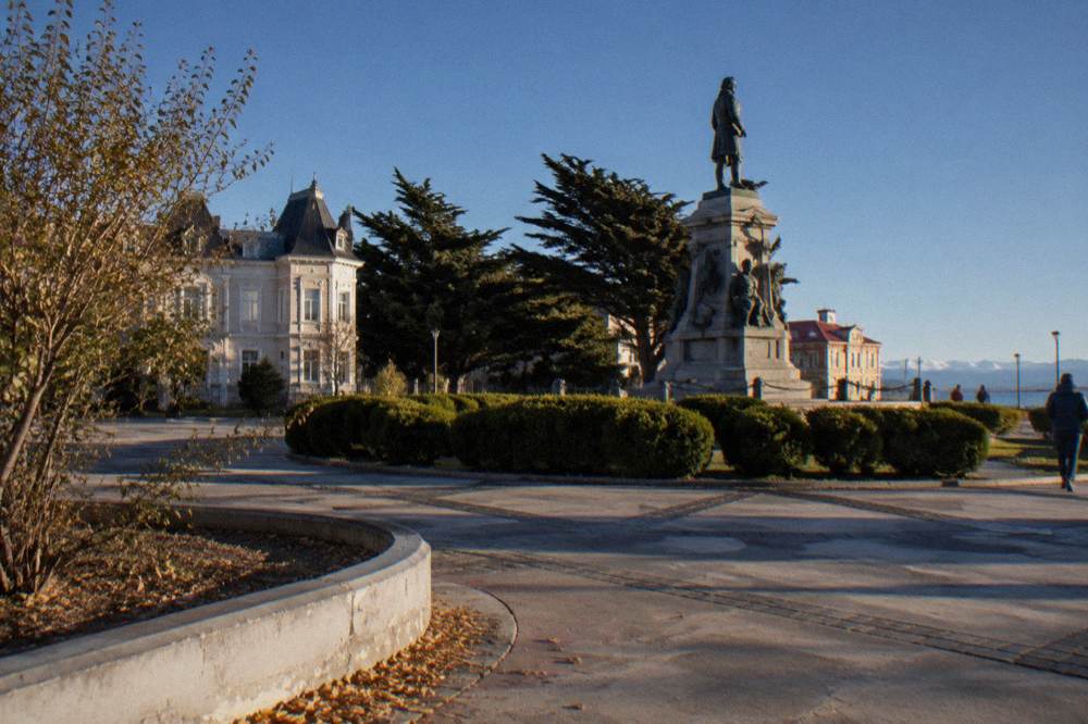 A monument in a plaza with colourful facades and distant hills under a wide sky