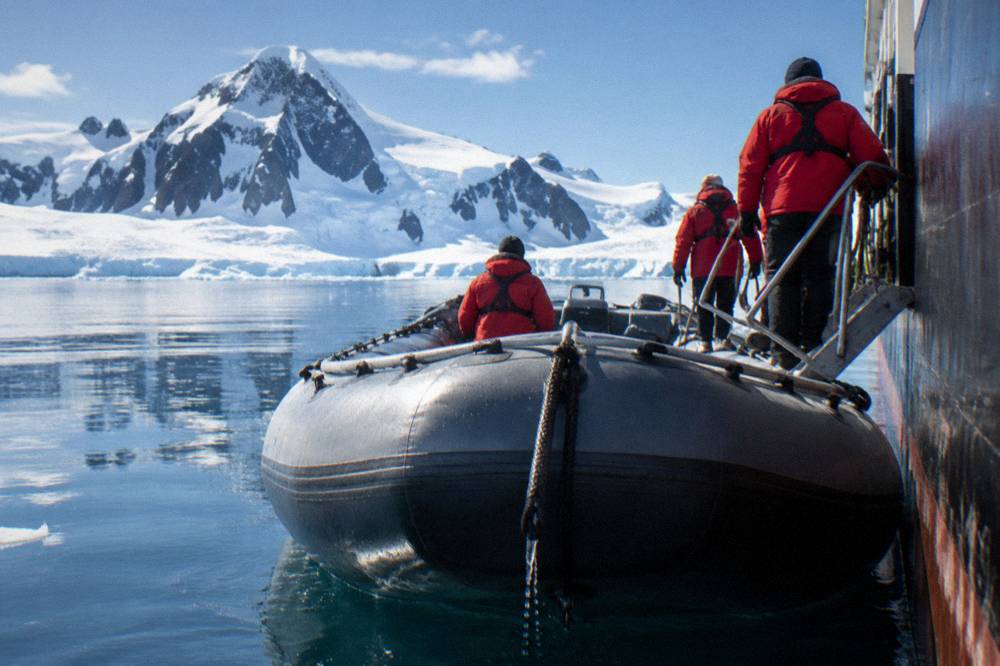Expedition guests in red parkas boarding Zodiacs from a ship gangway in calm polar water
