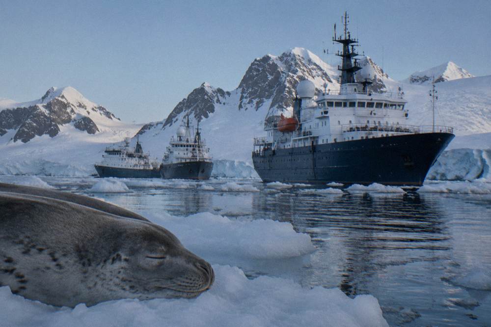 Two expedition ships at anchor in a sheltered Antarctic bay with snow-covered peaks behind