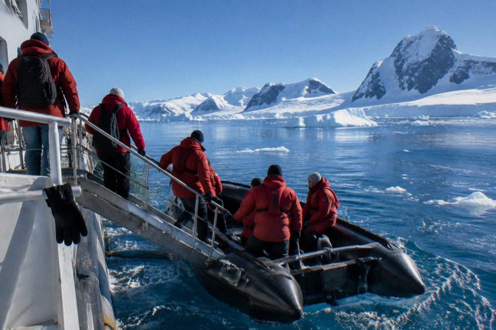 Passengers in red parkas boarding a Zodiac from an expedition ship gangway in icy water