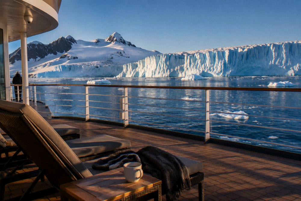 A luxury expedition ship's sundeck with loungers facing a wall of glacier ice under evening light