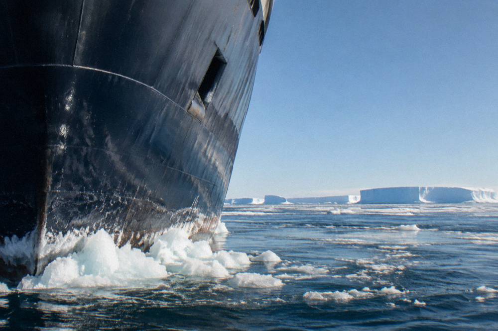 An expedition ship's bow cutting through brash ice under a bright polar sky
