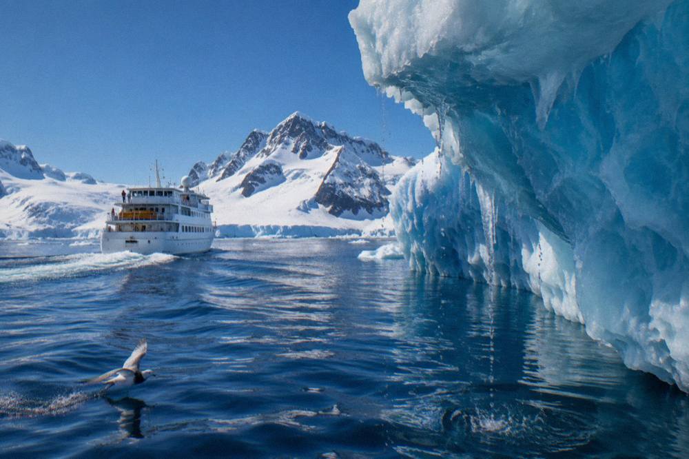 Small expedition ship cruising past a wall of blue glacier ice in the Antarctic Peninsula