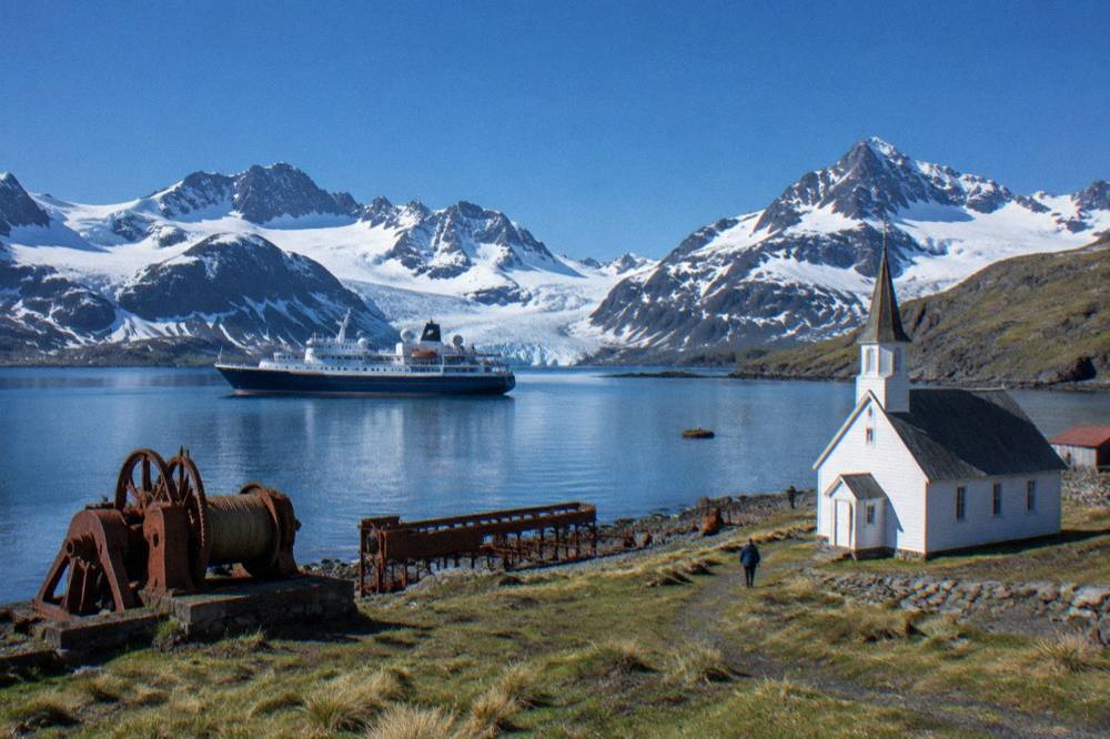 An expedition ship anchored in a sheltered bay with snow-covered mountains rising behind