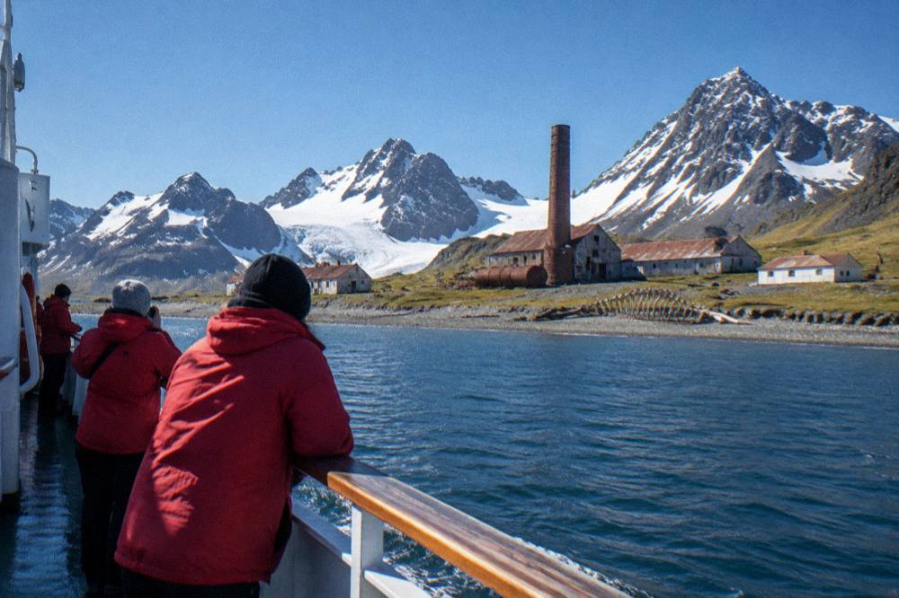 Red-jacketed travellers watching from a ship deck as jagged snowy peaks pass by