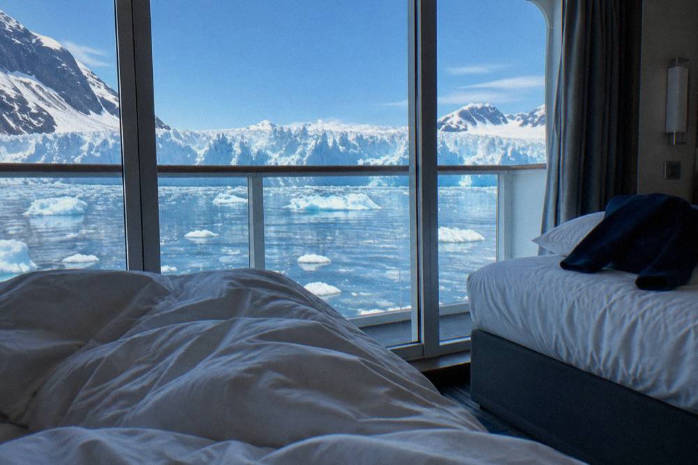 A balcony stateroom with twin beds and a view of Antarctic water through floor-to-ceiling glass