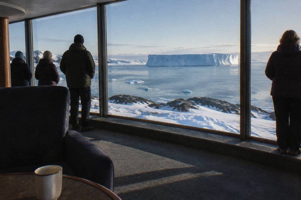 Guests in a glass-walled observation lounge watching icebergs pass during an Antarctic evening