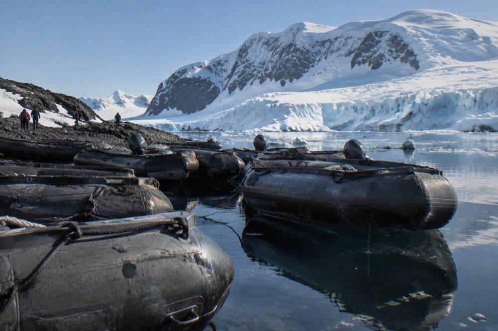 A fleet of Zodiacs lined up on a glassy Antarctic bay beneath a glacier face