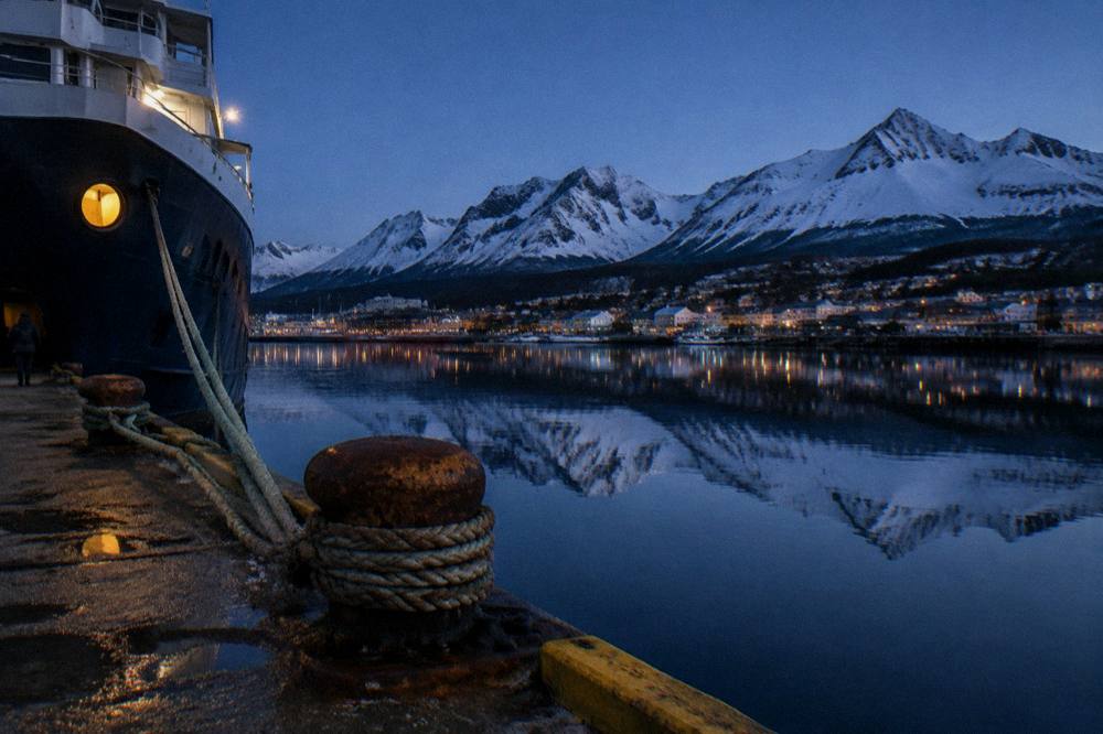 A quay with mooring lines and mountains reflected in still water at dusk