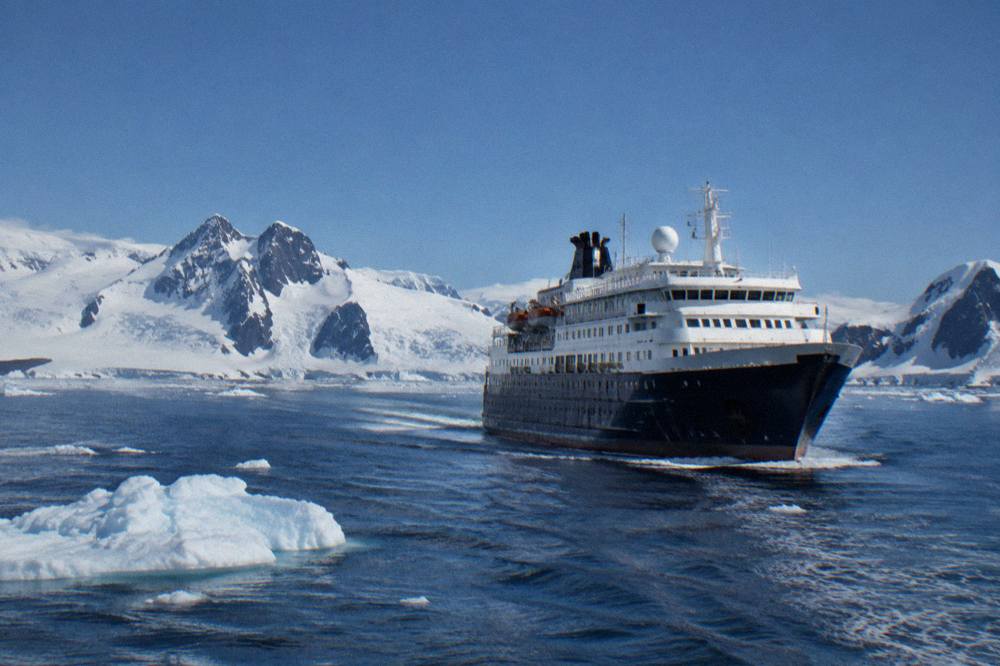 A modern expedition ship with a flat bow cruising through calm polar water scattered with small icebergs.