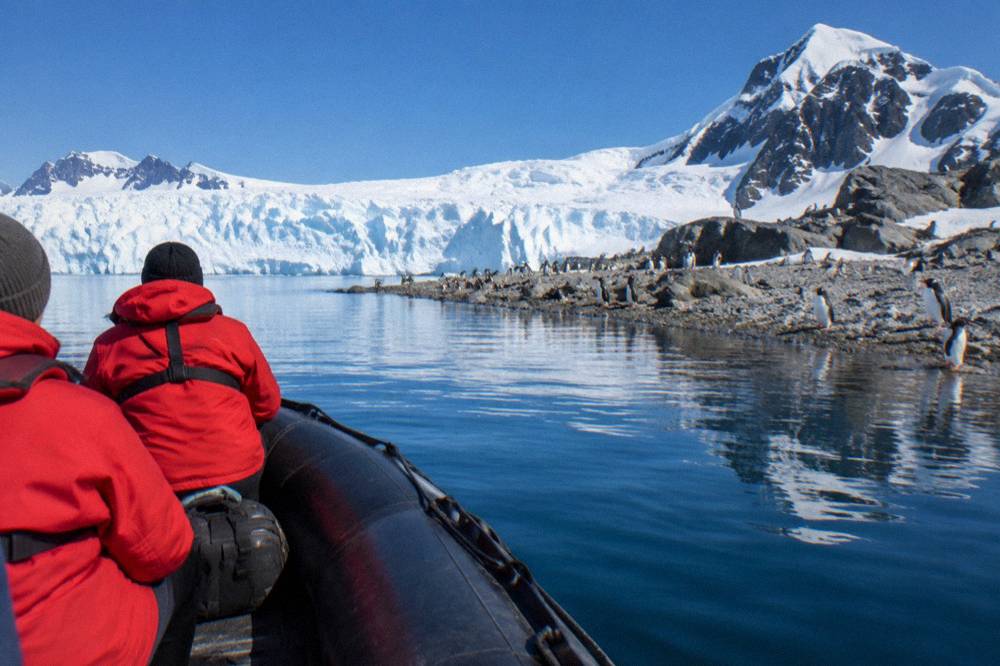 A zodiac boat carrying passengers in red parkas approaching a rocky Antarctic shoreline dotted with penguins.