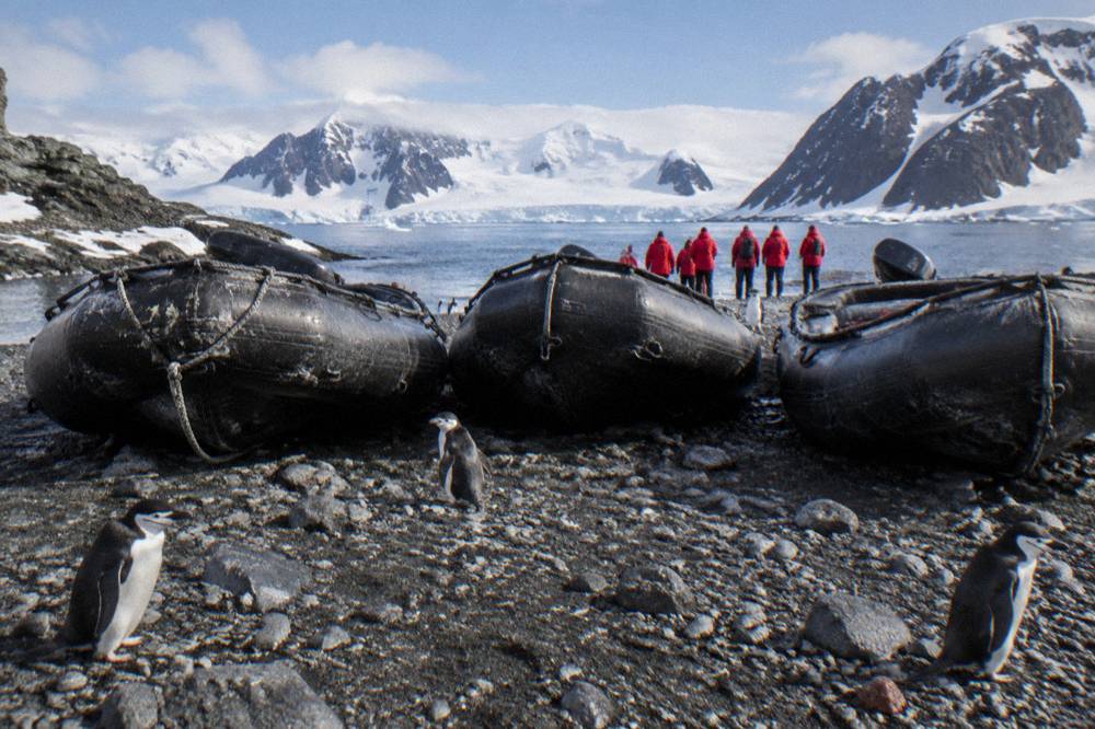 Zodiacs lined up on a rocky Antarctic beach with passengers in red parkas nearby