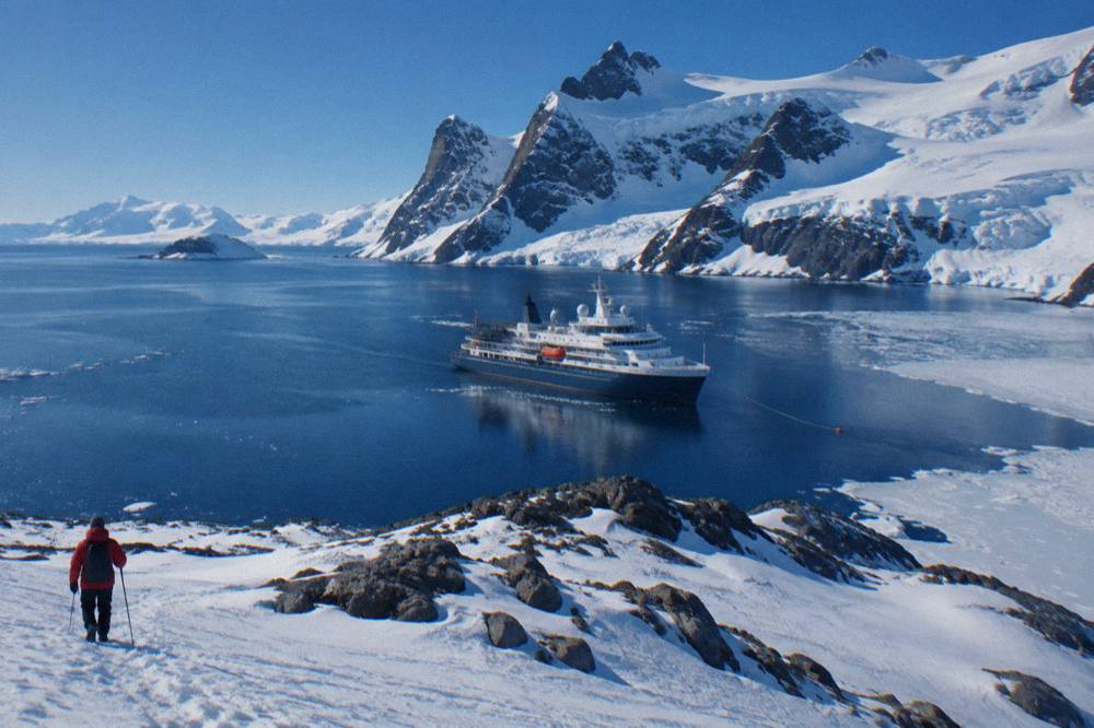 A modern expedition ship at anchor near a rocky Antarctic coastline with snow-covered peaks