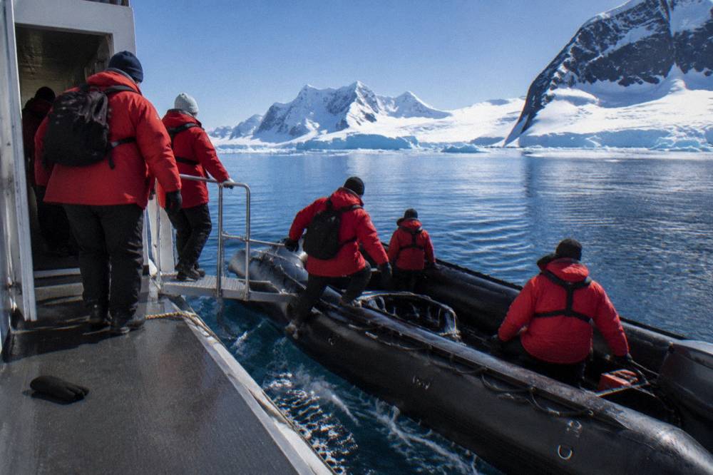 Passengers in red parkas boarding a Zodiac from an expedition ship gangway door in calm polar water