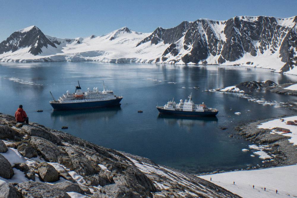 Two expedition ships moored in a calm Antarctic bay ringed by snow-covered mountains